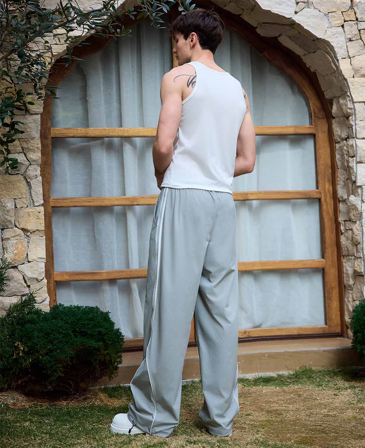 Man in light grey baggy wide leg sweatpants, white thick-soled shoes, back to camera, side stance on grass, showing loose fit and stripes.