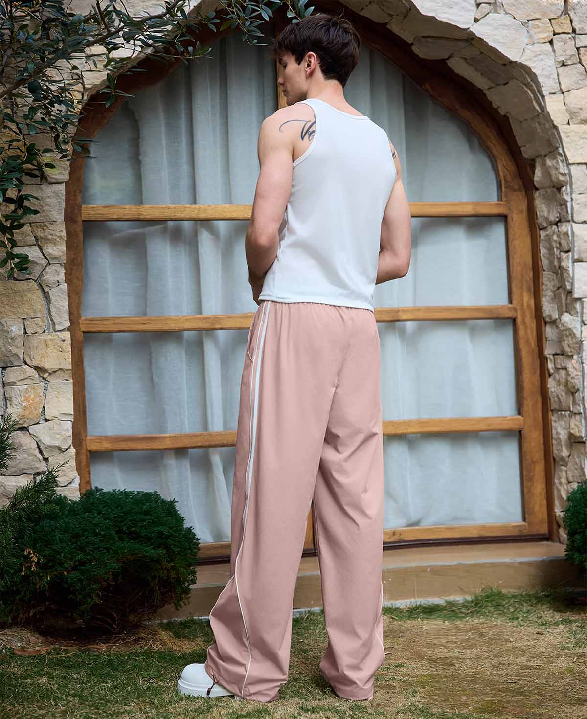 Man in pink baggy wide leg sweatpants, white thick-soled shoes, back to camera, side stance on grass, showing loose fit and stripes.