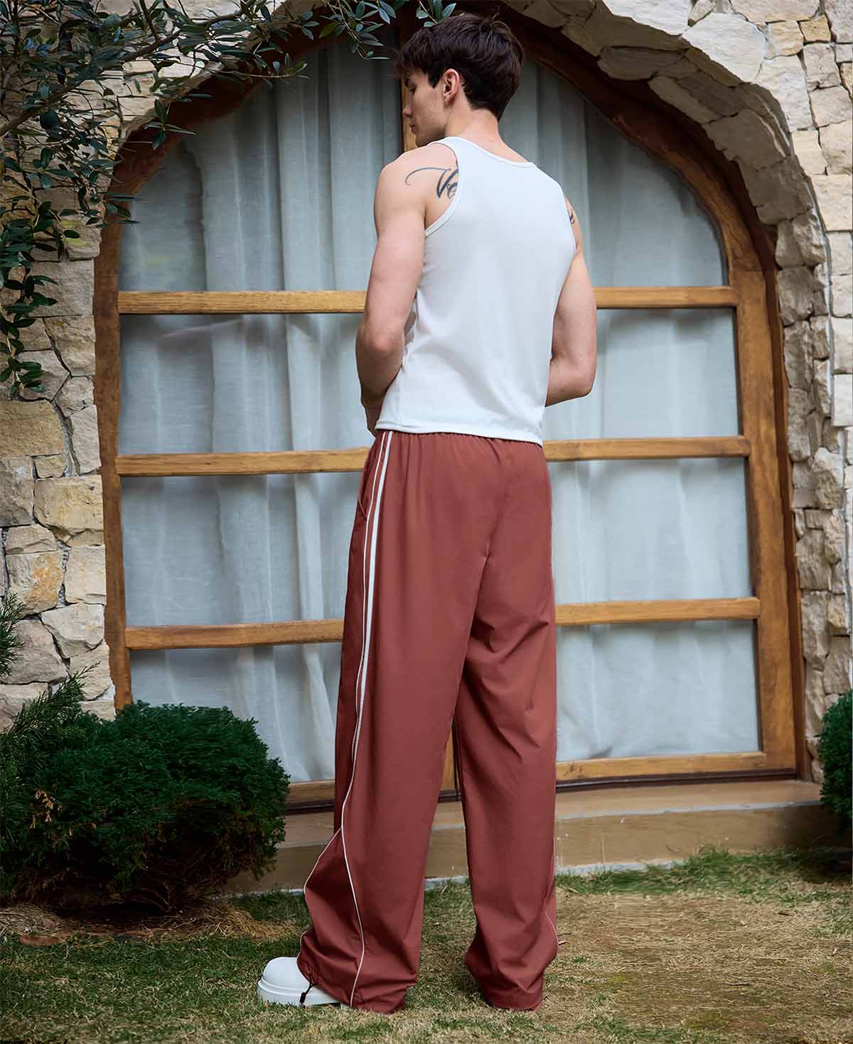 Man in red baggy wide leg sweatpants, white thick-soled shoes, back to camera, side stance on grass, showing loose fit and stripes.